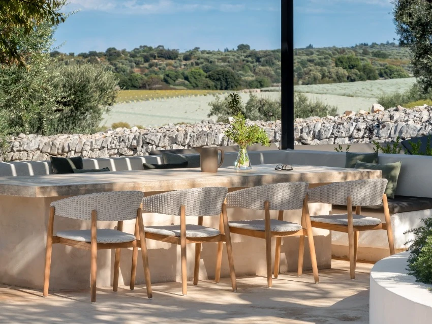 Mona dining chairs by Vincent Sheppard in Old Lace synthetic wicker with teak frames, arranged around a light stone outdoor table under a modern black pergola. 